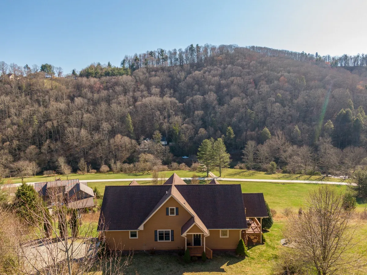 A broad exterior view of the property reveals the house nestled among mature trees with an expansive open field in the foreground. The spacious porch and surrounding landscape invite guests to connect with nature, perfect for outdoor gatherings or simply relaxing and enjoying wildlife.