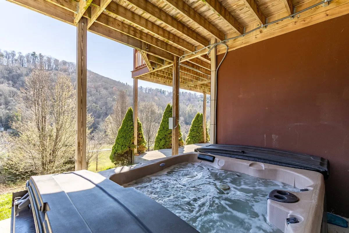 This exterior shot centers on the covered hot tub, set against a backdrop of rolling mountains and lush landscaping. Visible wooden beams give the space a rustic, inviting feel, while the bubbling hot tub promises year-round relaxation. The view from the hot tub stretches out to the mountains, emphasizing both comfort and natural beauty.