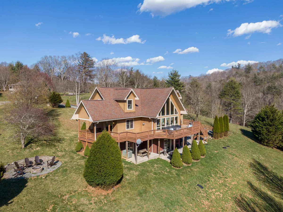 The image showcases the inviting exterior of a spacious mountain home with a large deck overlooking rolling hills, a cozy fire pit area surrounded by seating, and a beautifully landscaped yard perfect for outdoor activities. The setting is serene, nestled among trees with a backdrop of breathtaking mountain views, inviting guests to relax or gather for memorable evenings under the stars.