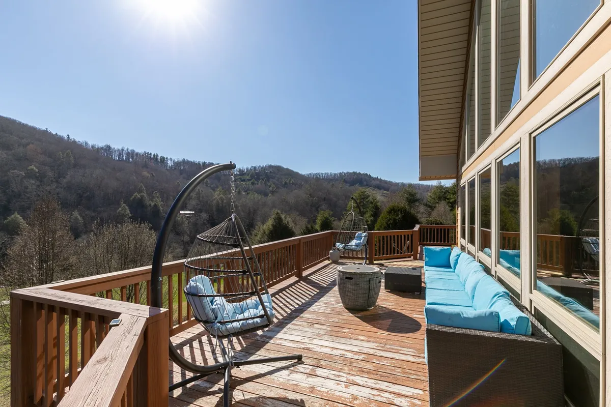 A cozy hanging chair sits on a wooden deck, accented with blue cushions and framed by sweeping mountain views. Large windows behind the deck hint at the indoor-outdoor connection, while the deck's vantage point emphasizes the breathtaking scenery and relaxation opportunities.