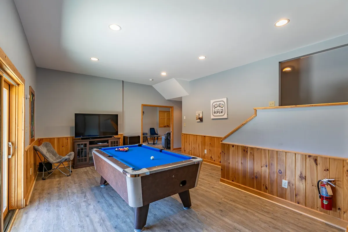 A lively interior scene featuring a pool table as the centerpiece, surrounded by comfortable seating and wood paneling. A television is mounted on the wall, and the atmosphere is casual and fun—perfect for group activities, watching movies, or relaxing after a game.