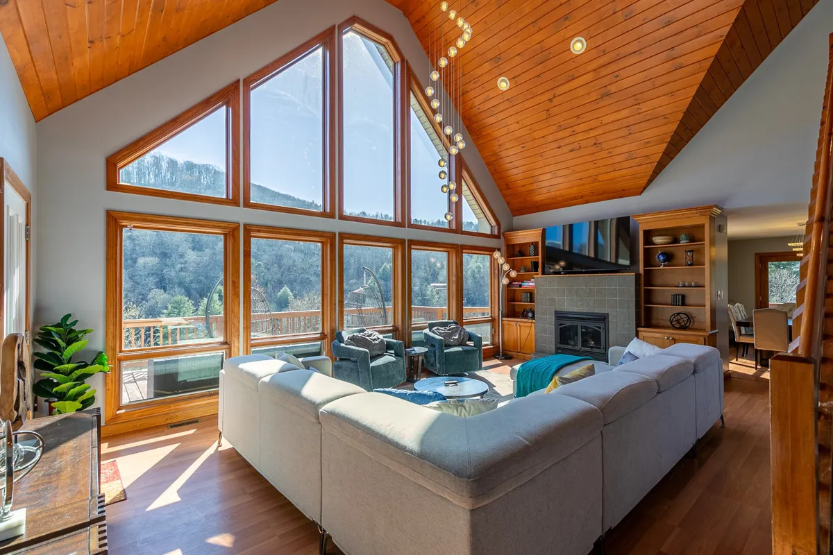 A welcoming living area with large windows that frame stunning mountain views, cozy modern furniture, and a glowing fireplace. The space is bathed in natural light, suggesting both the warmth of summer days and the comfort of winter nights. The deck just outside the windows hints at easy indoor-outdoor living.