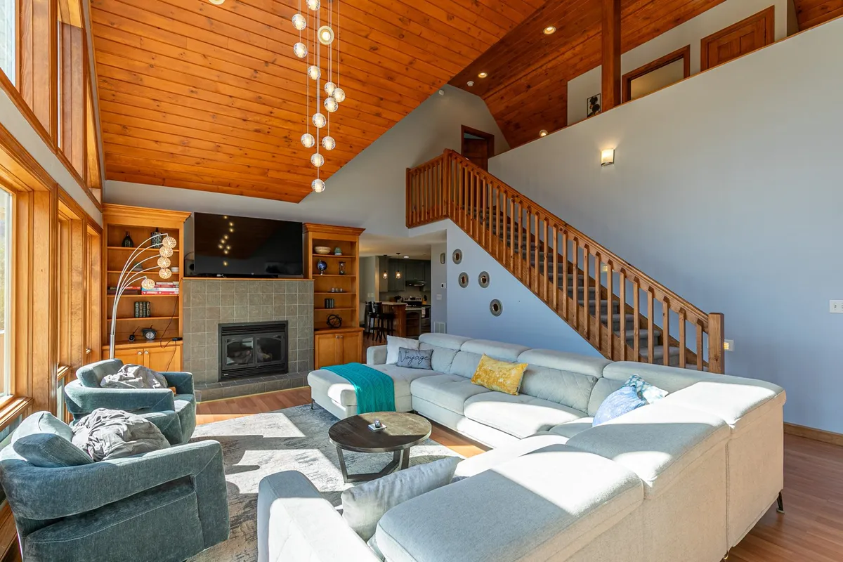 This image features a spacious great room with a large sectional sofa, a fireplace, a wooden staircase, and impressive floor-to-ceiling windows that allow natural light to flood the space. The modern light fixtures and warm wood tones create a cozy, inviting atmosphere, while the view hints at the surrounding landscape.
