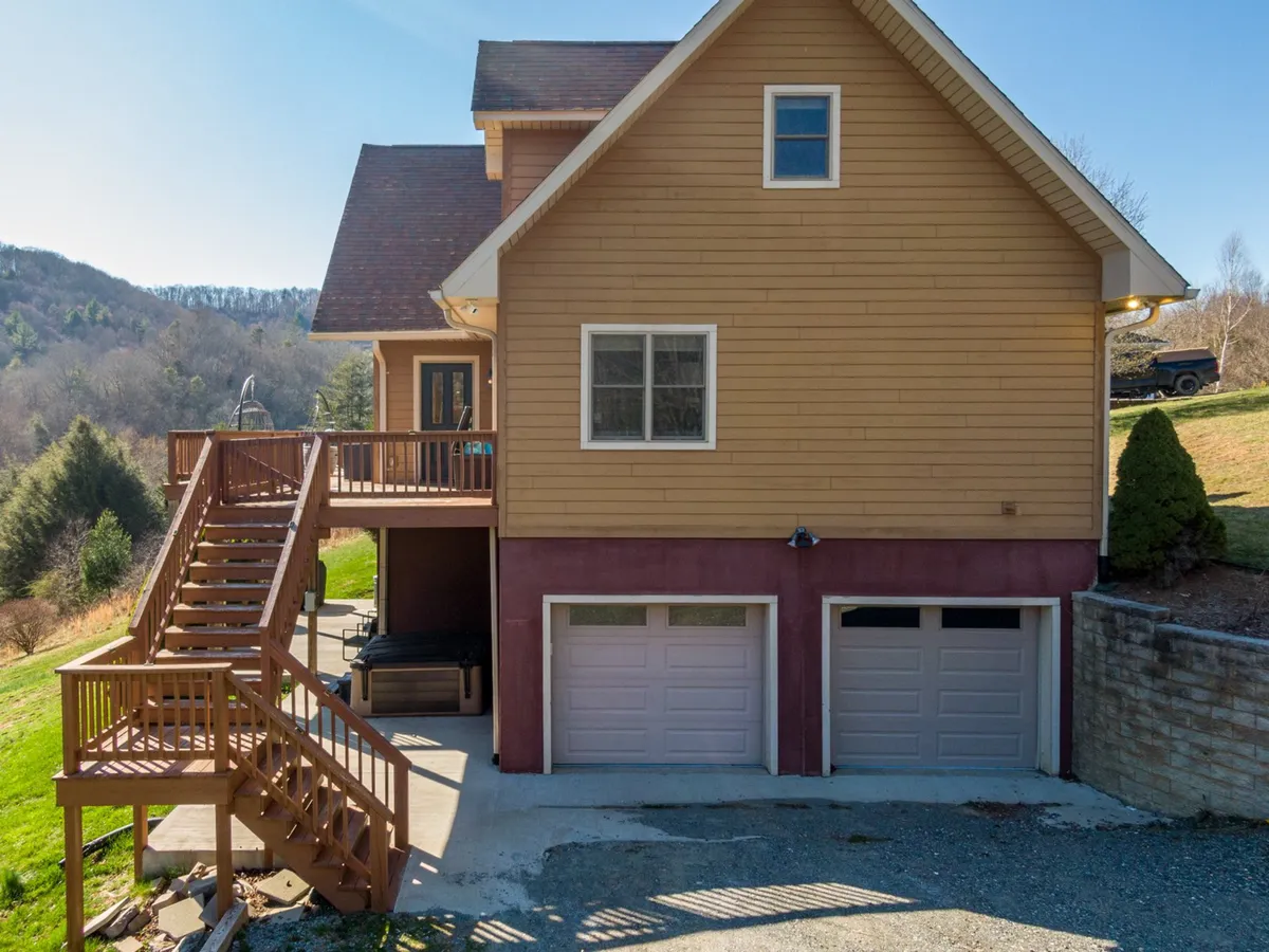 A staircase leads up to a spacious upper deck overlooking the surrounding landscape, with a two-car garage providing context for arrival and entry. The scene captures the tranquility of the home's exterior and the inviting nature of its outdoor spaces.