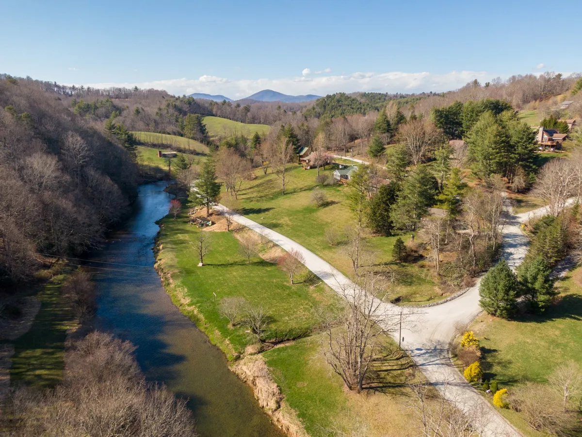 A tranquil riverside scene showing the South Fork of the New River winding through a lush green landscape, with mountains in the distance. The image includes a road, trees, and several houses nestled into the scenery, evoking the serene beauty of the Blue Ridge area.