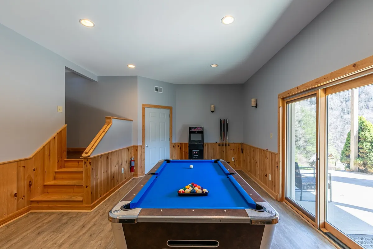 A vibrant, well-lit game room with a pool table, large windows letting in natural light, a wooden staircase, and a bar area. The space is ideal for group entertainment, family games, and unwinding after a busy day outdoors.